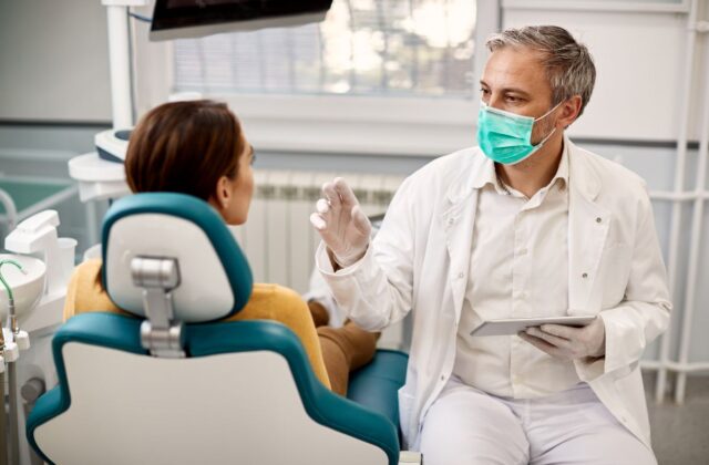 Dentist in a white coat and green mask explains treatment to a patient seated in a teal dental chair.